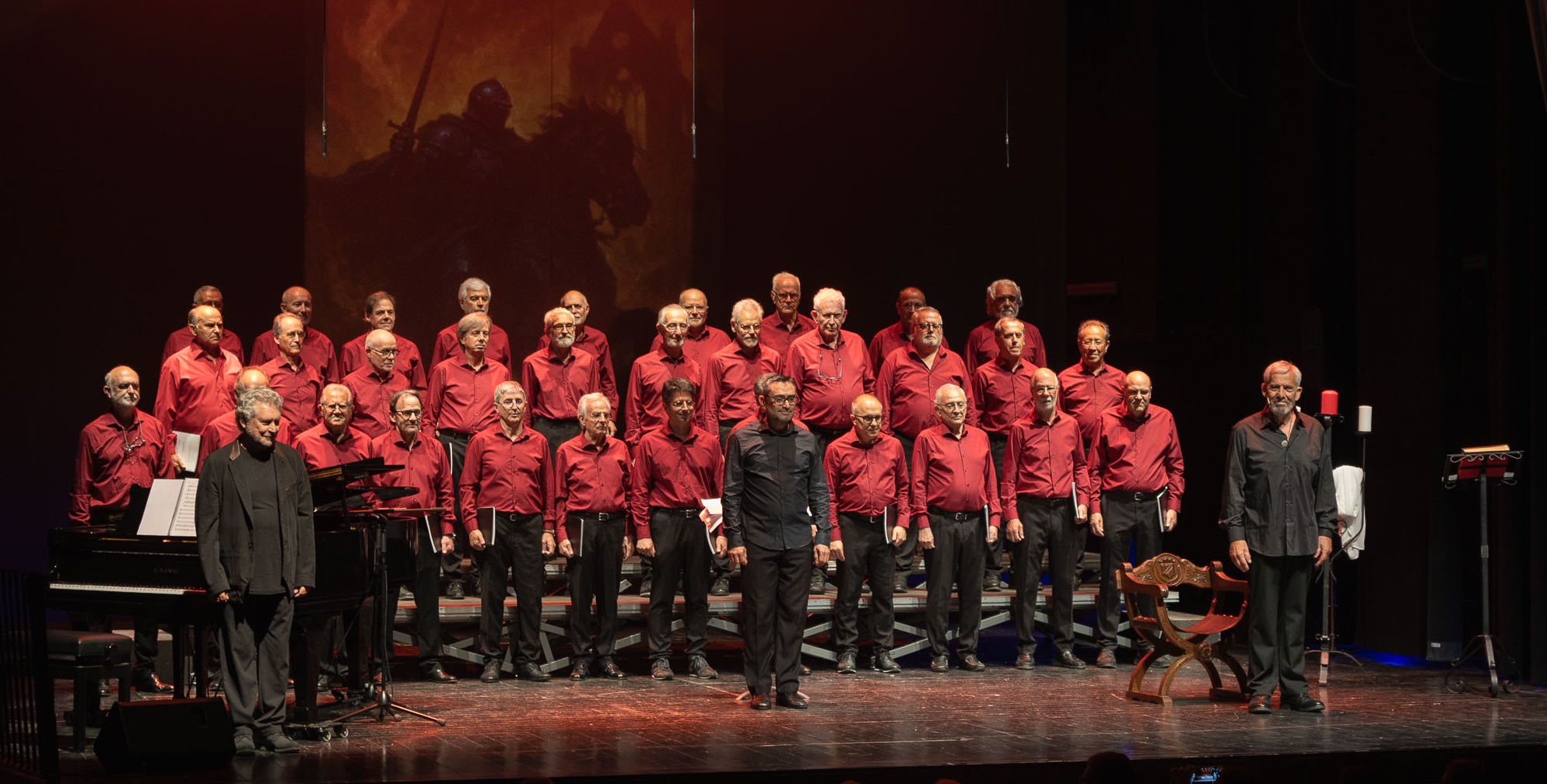 "Fotografia d'un cor format per homes amb camises vermelles actuant dalt d'un escenari. Estan situats sobre unes grades, acompanyats d'un piano de cua a l'esquerra i amb un gran teló de fons que mostra la imatge d'un cavaller a les fosques.